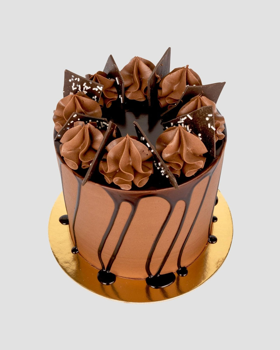 The Chocolate Lovers Cake by Jenna Rae Cakes is a tall, round cake with smooth frosting, chocolate drip, chocolate cream swirls, chocolate shards, and white sprinkles on a gold board against a plain background.