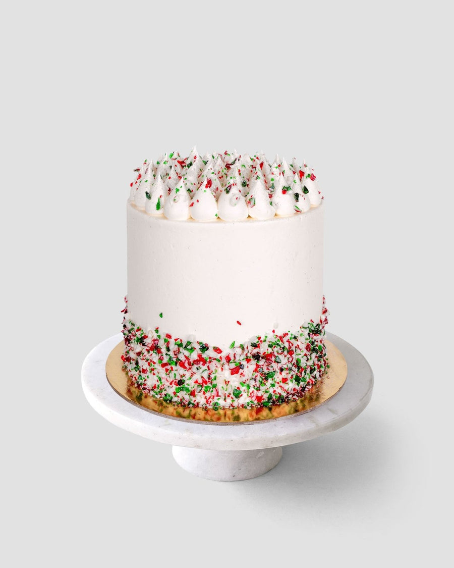 The Candy Cane Cake by Jenna Rae Cakes is a tall, round cake with smooth white frosting and red and green sprinkles on the bottom and top, shown on a white marble cake stand against a light gray background.