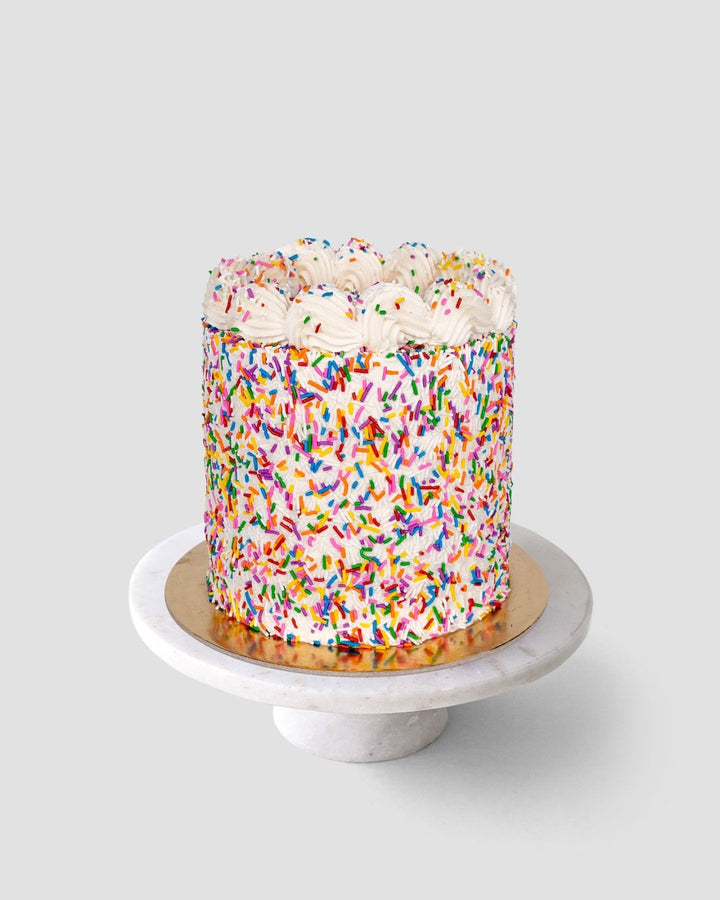 The Last Minute Cakes by Jenna Rae Cakes features a round cake covered in rainbow sprinkles and swirls of white frosting, set on a gold cake board atop a white marble stand against a light gray background.