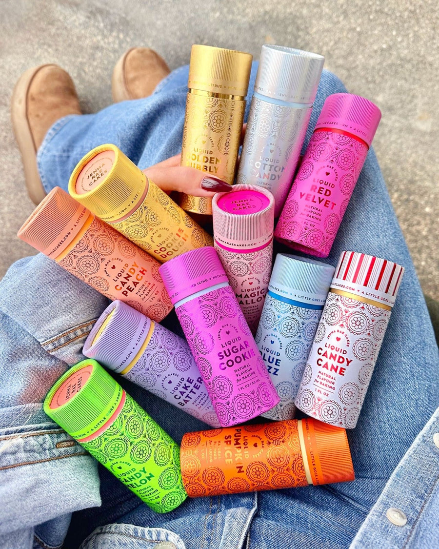A person wearing blue jeans holds a pack of Jenna Rae Cakes Liquid Cookie Dough (6-pack), each tube featuring unique flavors and designs. Tan shoes are visible in the background.