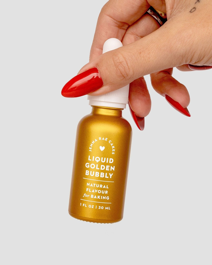 A hand with long red nails holds a small gold bottle of Jenna Rae Cakes Liquid Golden Bubbly - Package of 6, Natural Flavour for Baking, 1 FL OZ / 30 mL, against a plain light background.