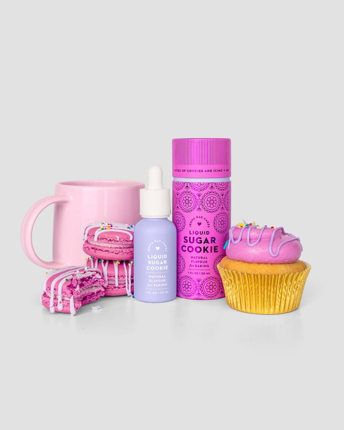 Jenna Rae Cakes’ Liquid Sugar Cookie: a pastel purple serum bottle with dropper, paired with a pink mug, tall pink spray can, pink macaron, broken sprinkled cookie, and pink frosted cupcake—all set against a light gray background.