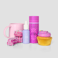 Jenna Rae Cakes’ Liquid Sugar Cookie: a pastel purple serum bottle with dropper, paired with a pink mug, tall pink spray can, pink macaron, broken sprinkled cookie, and pink frosted cupcake—all set against a light gray background.