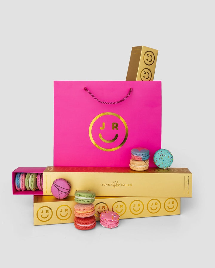 A bright pink Jenna Rae Cakes gift bag with a gold smiley face logo sits among yellow Macaron Gift Boxes filled with colorful macarons, some stacked and some scattered, all set against a white background.
