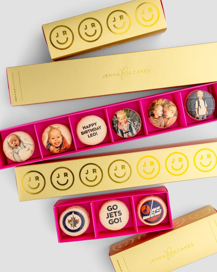 The Macaron Message Gift Box from Jenna Rae Cakes features rectangular yellow boxes with custom macarons displaying smiling faces, children’s photos, messages like HAPPY BIRTHDAY LEO!, GO JETS GO!, and Winnipeg Jets logos.