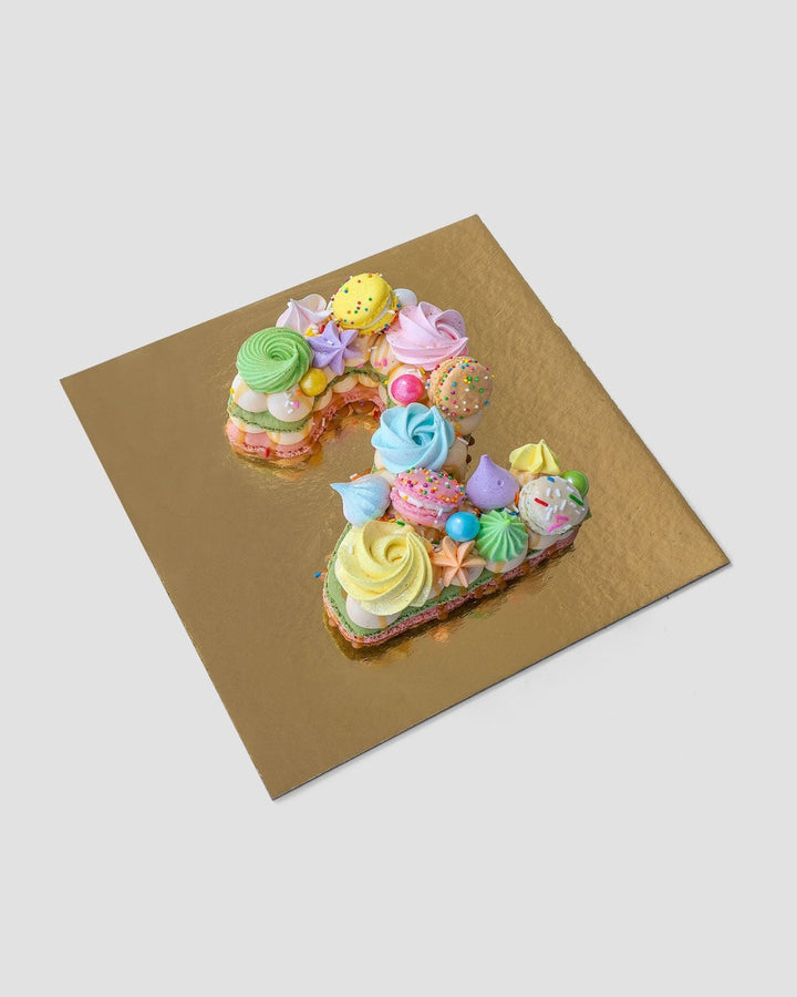 The Jenna Rae Cakes Macaron Number Cake, shaped as the number 2 and topped with pastel meringues, sprinkles, and candies, is presented on a square gold board against a light gray background.
