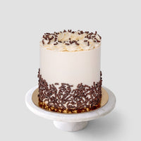 The Marble Cake by Jenna Rae Cakes features smooth white frosting, piped swirls, and chocolate shavings around the base. It sits on a white marble cake stand against a simple light background.