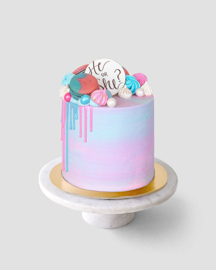 The Reveal Works Cake by Shop Jenna Rae Cakes features pastel pink and blue frosting with dripping icing, colorful meringues, and a “He or She?” white chocolate disk, all presented on a marble cake stand against a gray background.
