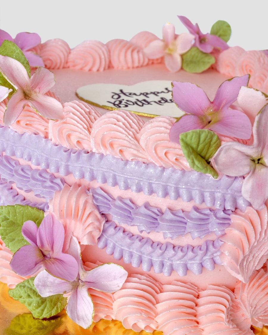 The Sugar Flower Heart Cake by Jenna Rae Cakes is a heart-shaped cake with pink and purple buttercream swirls, topped with matching flowers and a white heart-shaped Happy Birthday plaque.