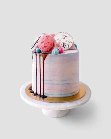 The Half Works Cake by Jenna Rae Cakes is a pastel birthday cake topped with a pink macaron, blue and pink candies, chocolate drizzle, and a white chocolate Happy 17th Birthday disc on a marble stand.