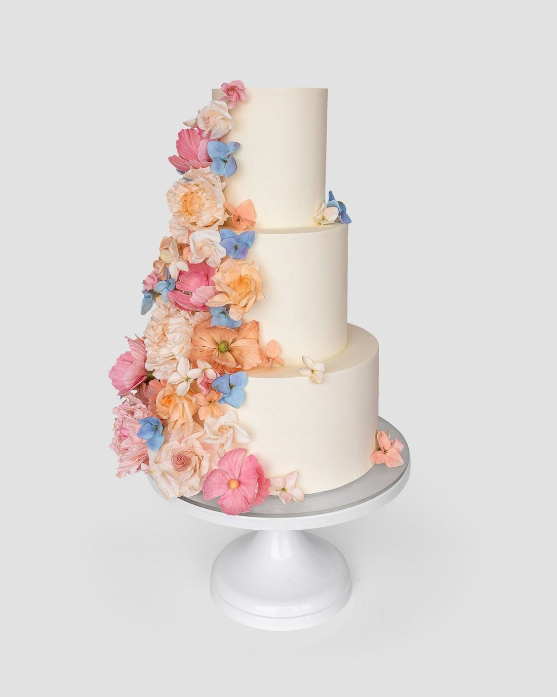 Jenna Rae Cakes Wedding Cakes ($1,000+) feature three tiers in white, adorned with cascading pastel flowers—pink, peach, and blue—presented on a white stand against a simple light background.