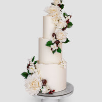 A three-tiered white cake decorated with white flowers, green leaves, and small red berries, displayed on a white cake stand against a plain background.