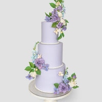 A three-tiered lavender Wedding Cake by Jenna Rae Cakes ($1,000+) features lifelike purple, blue, and white sugar flowers with green leaves, presented on a white cake stand against a simple light backdrop.