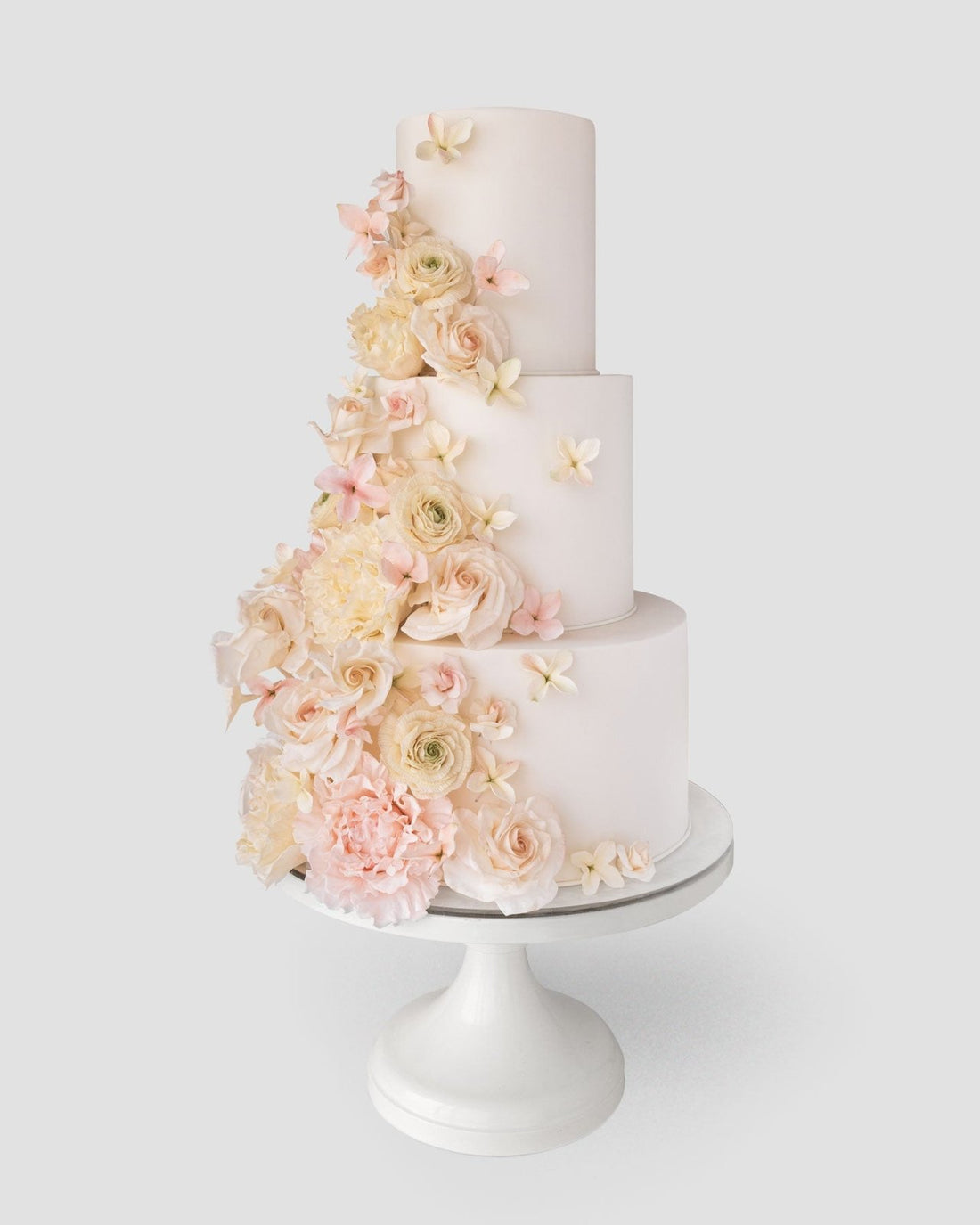 The Jenna Rae Cakes Wedding Cake ($1,000+) is a three-tiered white cake with cascading pastel roses and peonies, displayed on a white stand against a plain light background.
