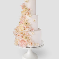 The Jenna Rae Cakes Wedding Cake ($1,000+) is a three-tiered white cake with cascading pastel roses and peonies, displayed on a white stand against a plain light background.