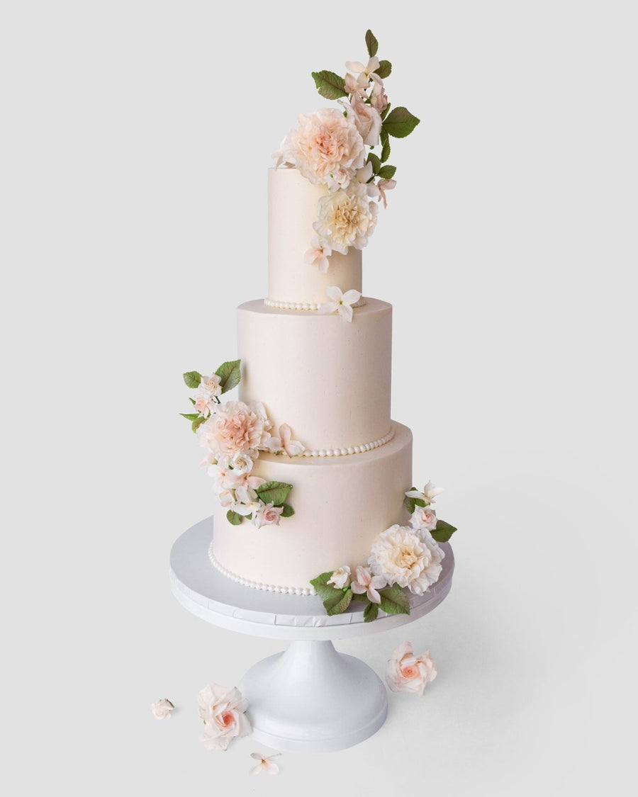 A three-tiered white Wedding Cake by Jenna Rae Cakes ($1,000+) features lifelike pale pink and white flowers with green leaves, presented on a white cake stand with additional flowers arranged at the base.