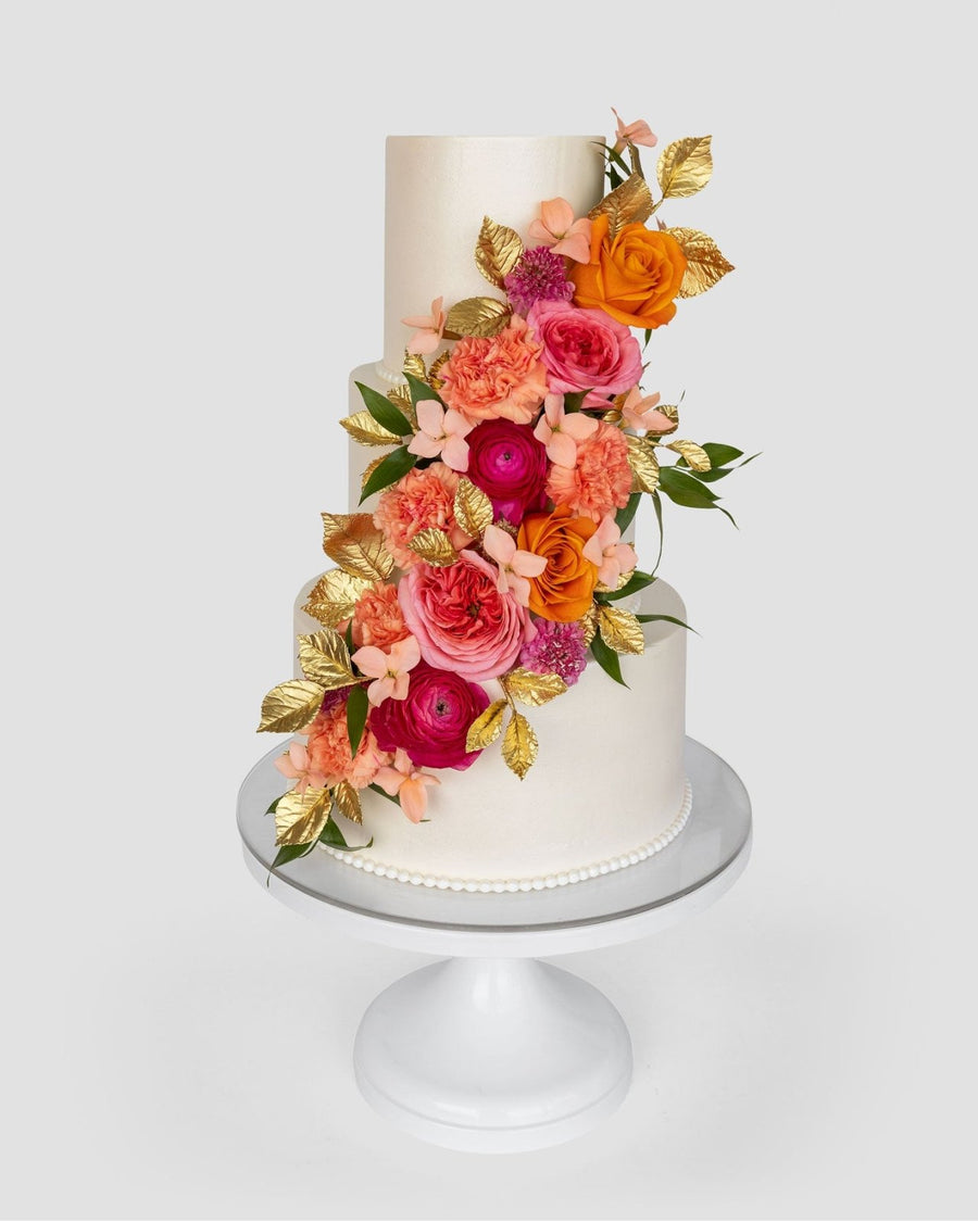 Three-tiered white cake decorated with vibrant pink, orange, and peach flowers, green leaves, and gold-painted leaves cascading down the side. The cake sits on a white pedestal stand against a light background.