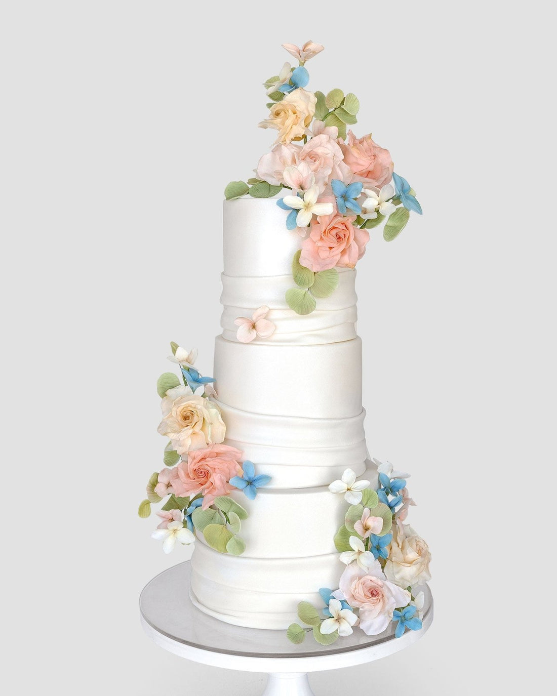The Wedding Cakes $1,000+ by Jenna Rae Cakes features a three-tiered white cake adorned with pastel pink, peach, blue, and white flowers plus green leaves, displayed on a white stand against a simple backdrop.