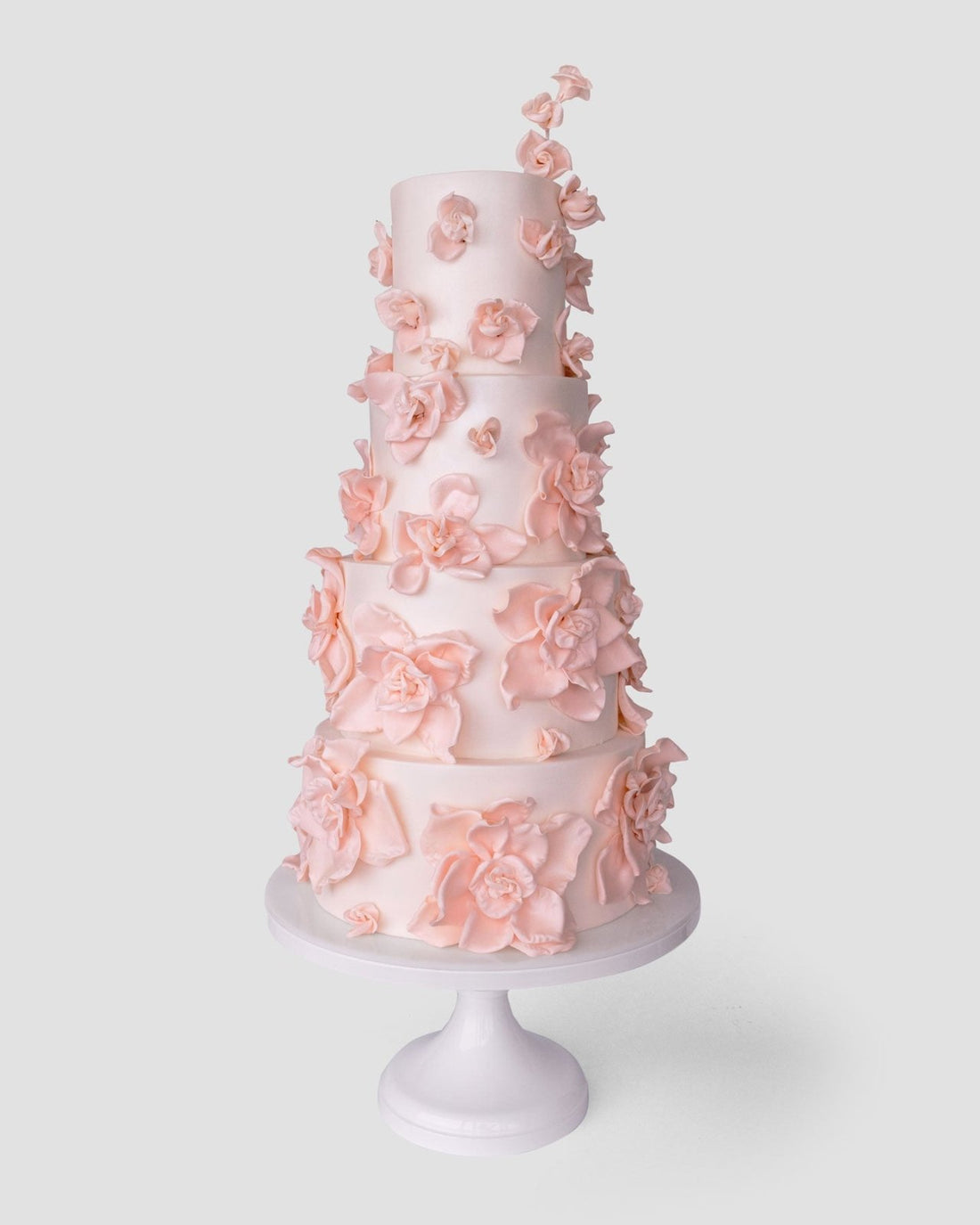 A tall, five-tier white wedding cake by Jenna Rae Cakes ($1,000+) features delicate light pink sugar flowers and is displayed on a white stand against a plain light background.
