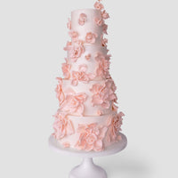 A tall, five-tier white wedding cake by Jenna Rae Cakes ($1,000+) features delicate light pink sugar flowers and is displayed on a white stand against a plain light background.