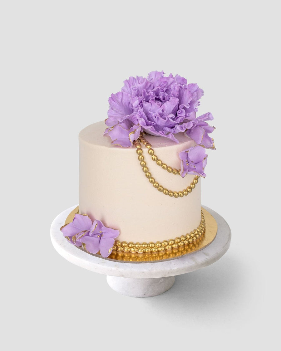 A beige-frosted cake from Jenna Rae Cakes’ Wedding Cakes Under $200, set on a white stand and adorned with gold beads and large purple sugar flowers on top and the side.