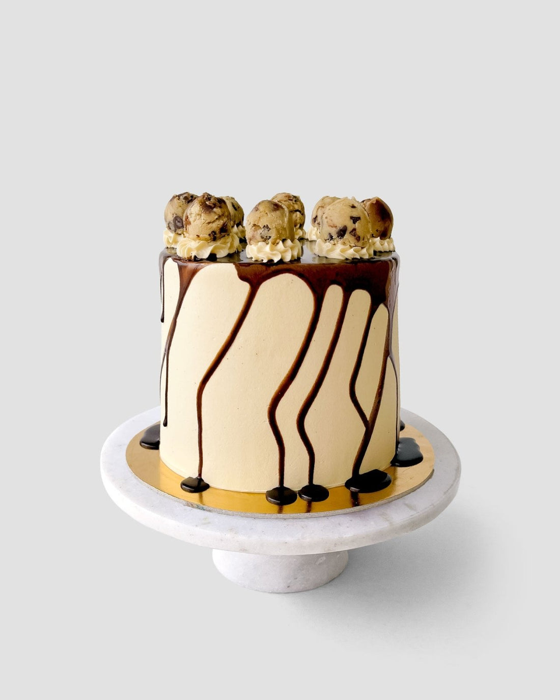 The Cookie Dough Cake by Jenna Rae Cakes features smooth beige frosting, chocolate drips down the sides, and scoops of cookie dough on top, displayed on a marble cake stand against a plain background.