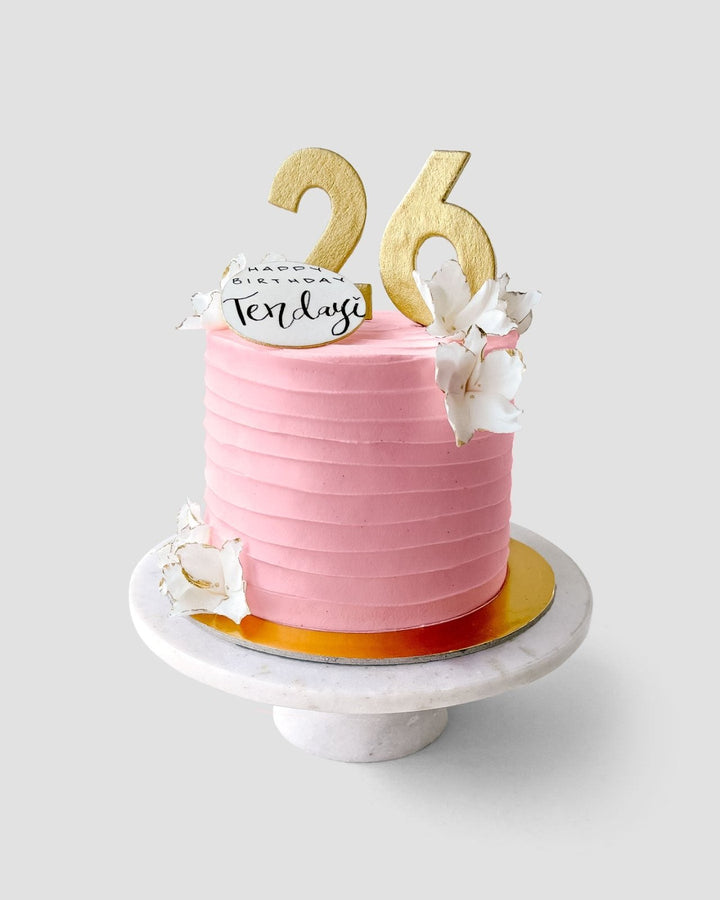 The Design Your Own Cake by Jenna Rae Cakes features pink frosting, gold 26 toppers, white flower decorations, and a Happy Birthday Tenley card, displayed on a marble cake stand.