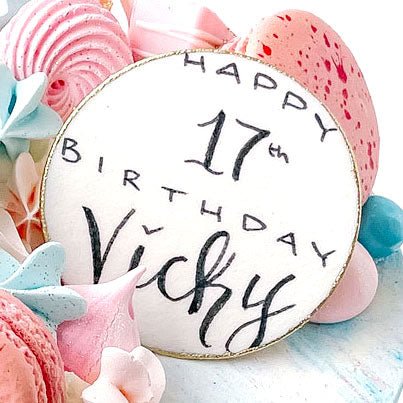 The Greeting Plaque by Shop Jenna Rae Cakes is a round white cookie with Happy 17th Birthday Vicky in black icing, surrounded by colorful meringues and pink, blue, and white macarons.