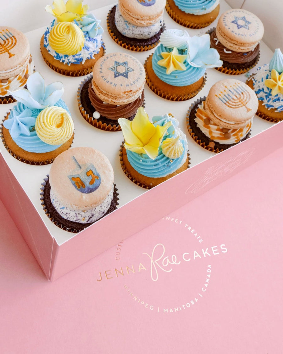 Hanukkah Works Cupcakes by Jenna Rae Cakes: a box of Hanukkah-themed cupcakes and macarons with blue, yellow, and white frosting, decorated with stars, menorahs, and dreidels. Logo displayed on a pink surface.