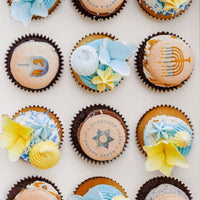 Hanukkah Works Cupcakes by Jenna Rae Cakes are decorated with blue, yellow, and white flowers, menorahs, dreidels, and Stars of David—perfectly arranged for a festive Hanukkah celebration.