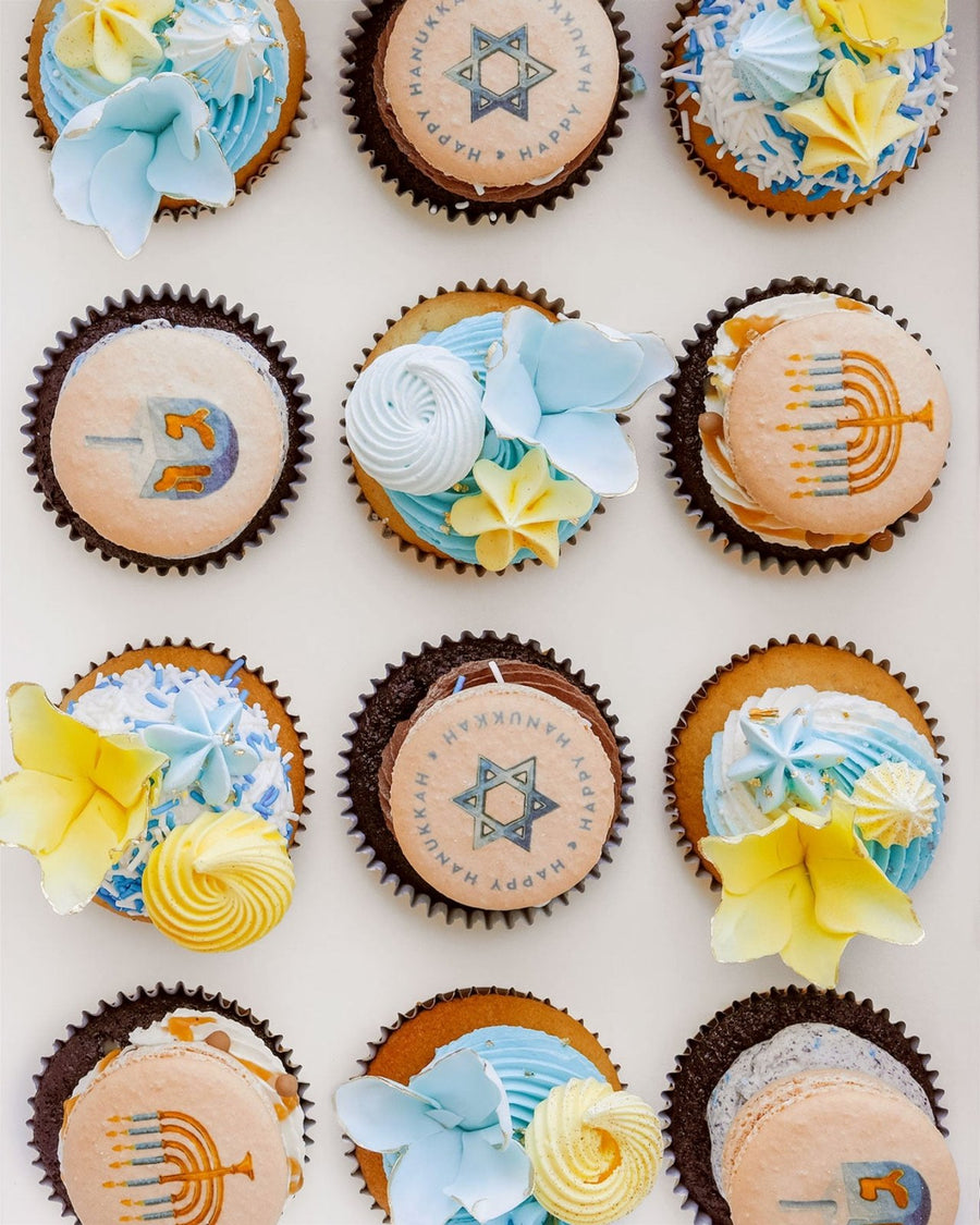 Hanukkah Works Cupcakes by Jenna Rae Cakes are decorated with blue, yellow, and white flowers, menorahs, dreidels, and Stars of David—perfectly arranged for a festive Hanukkah celebration.