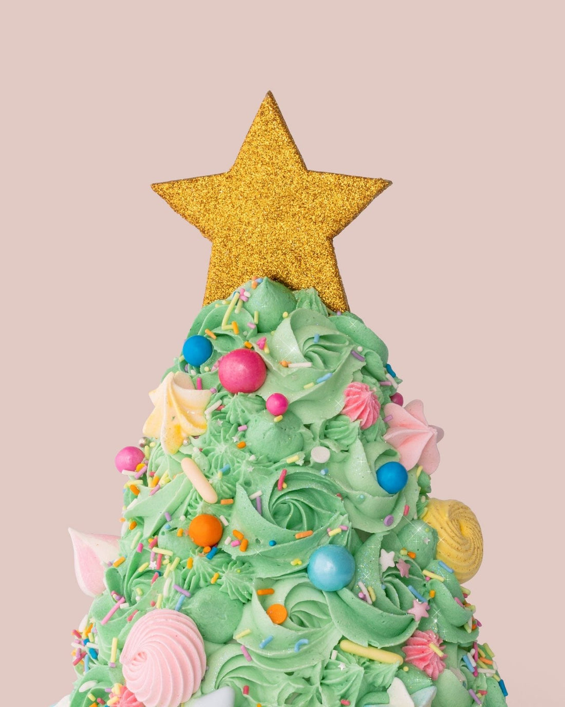 The Christmas Tree Cake by Jenna Rae Cakes features green frosting, colorful candy ornaments, and a glittery gold star on top, set against a plain light pink background.