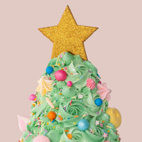 The Christmas Tree Cake by Jenna Rae Cakes features green frosting, colorful candy ornaments, and a glittery gold star on top, set against a plain light pink background.