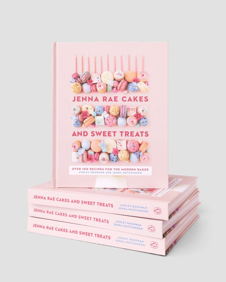 A stack of four Jenna Rae Cakes and Sweet Treats Cookbooks by Jenna Rae Cakes, with one standing upright on top, displays colorful macarons and desserts on the pink covers against a light grey background.