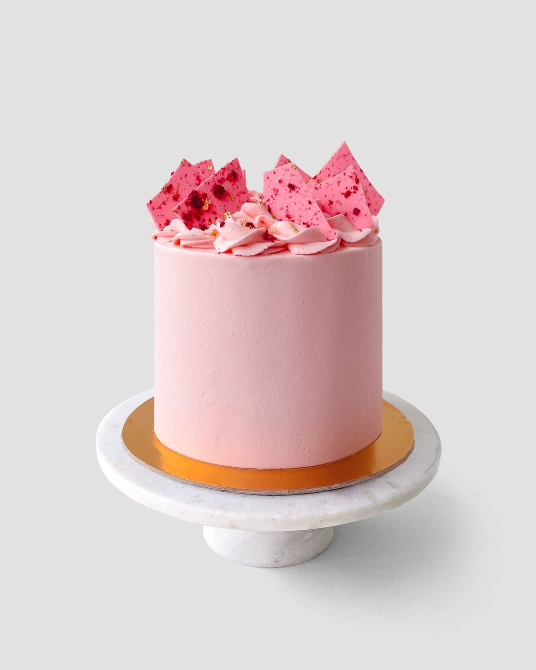 The Lemon Raspberry Cake by Jenna Rae Cakes features smooth pink frosting, pink shards, and swirls, displayed on a white marble cake stand against a plain light background.