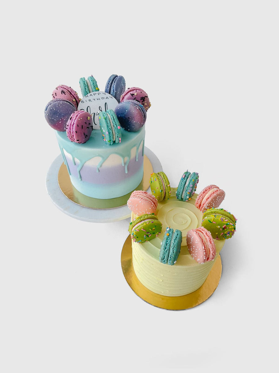 The Macaron Crown Cake by Jenna Rae Cakes features two cakes topped with colorful macarons, one with pastel blue and purple icing and white drip, the other with light yellow icing. Both sit on gold bases against a plain white background.