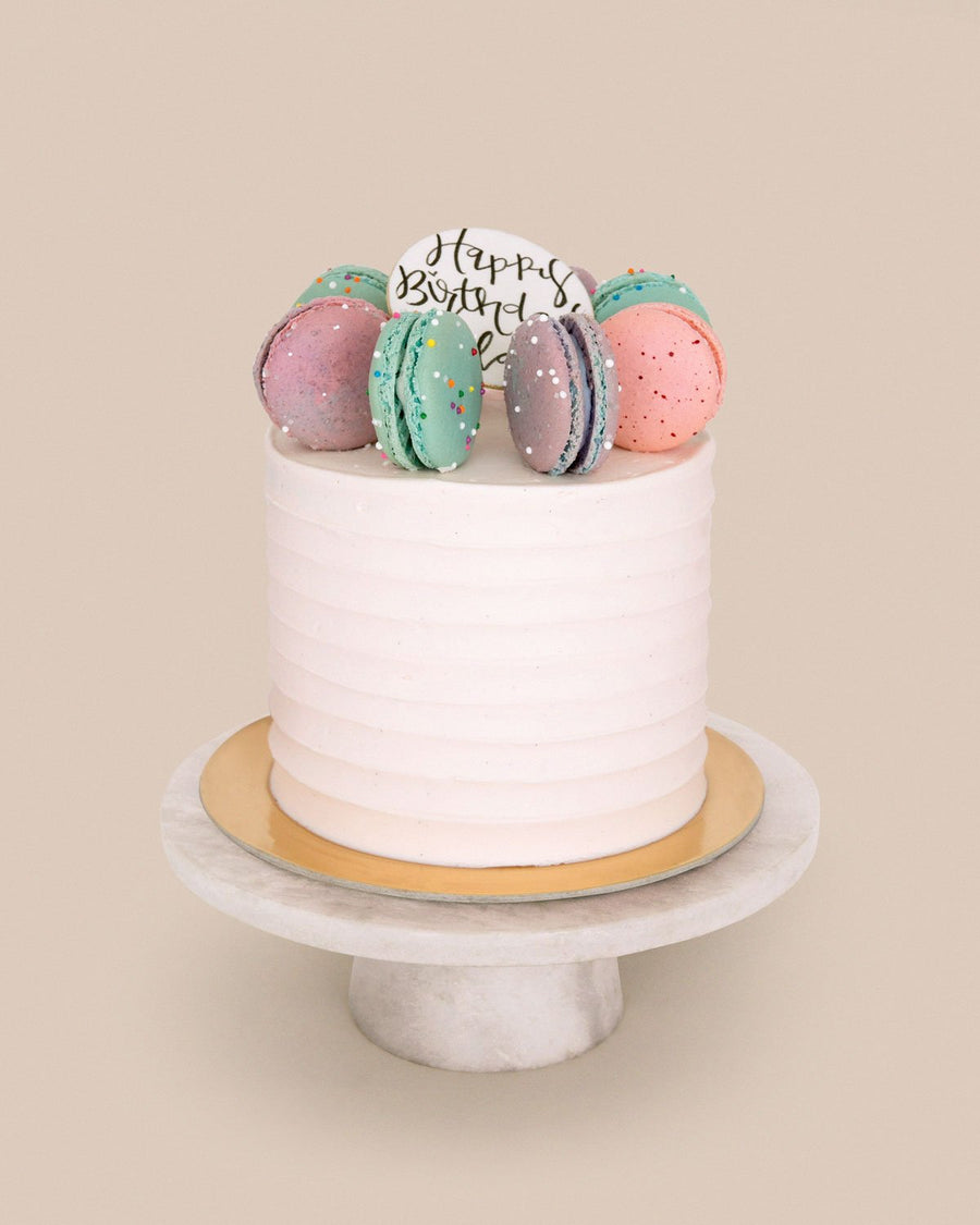 The Macaron Crown Cake by Jenna Rae Cakes features white frosting, colorful macarons, and a Happy Birthday sign, displayed on a round marble cake stand against a beige background.