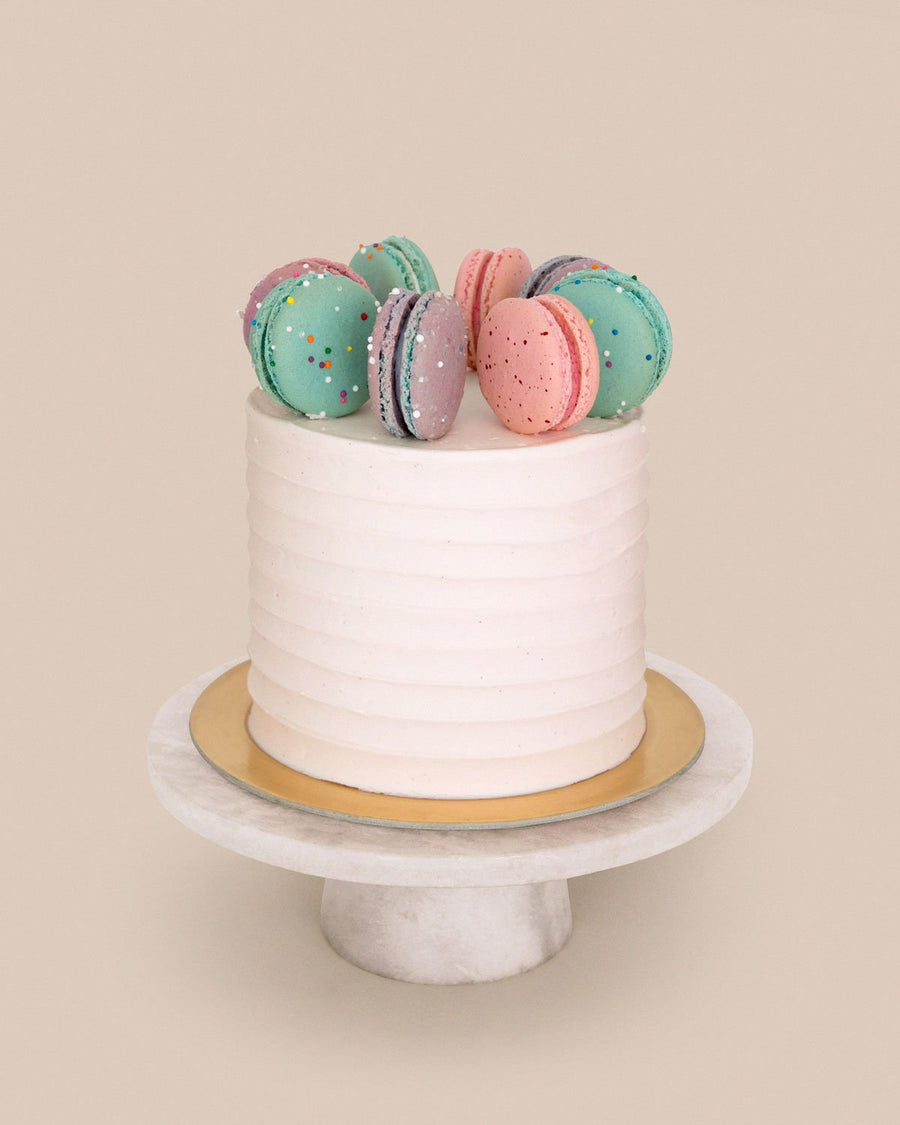 The Macaron Crown Cake by Jenna Rae Cakes features a white frosted finish topped with colorful macarons, displayed on a gold cake board and white marble stand, set against a plain beige background.