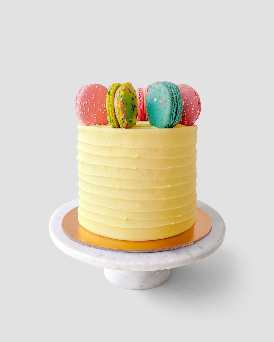 The Macaron Crown Cake by Jenna Rae Cakes features a yellow frosted finish with textured sides and five colorful macarons on top, displayed on a white marble cake stand against a plain light gray background.