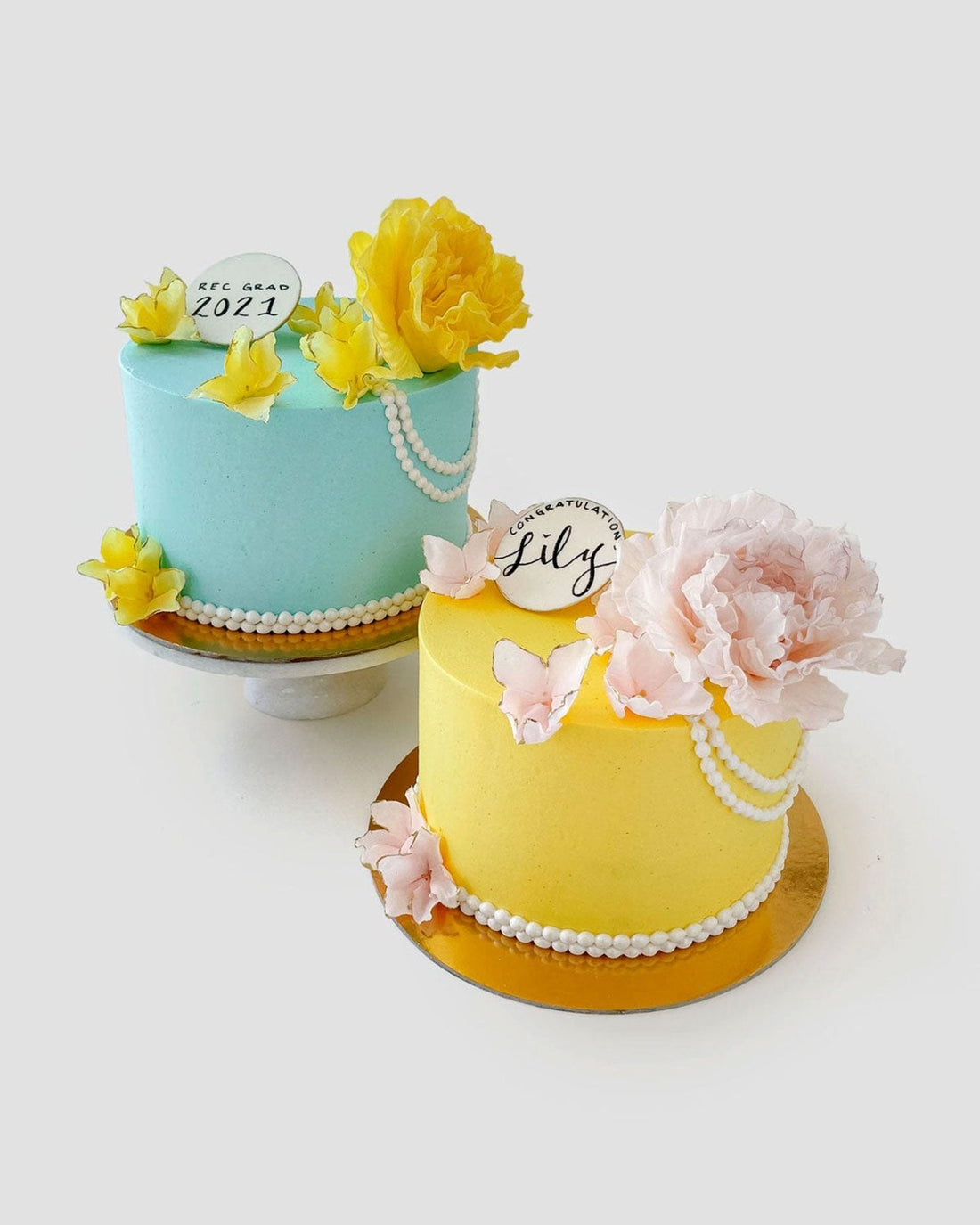 Two Jenna Rae Cakes Peony and Pearls Cakes: one blue with yellow flowers, pearls, and a 2021 topper; the other yellow with pink flowers, pearls, and a Congratulations Lily sign—both on gold cake boards against a white background.