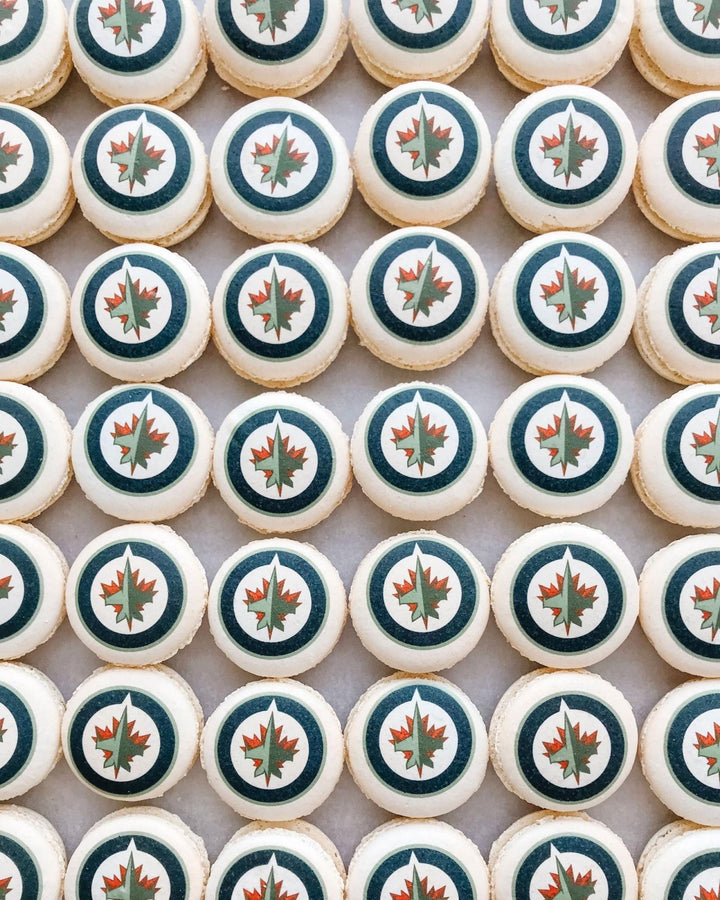 A grid of Printed Macarons by Jenna Rae Cakes, each topped with the Winnipeg Jets logo featuring a red maple leaf and jet in a blue and white circle.