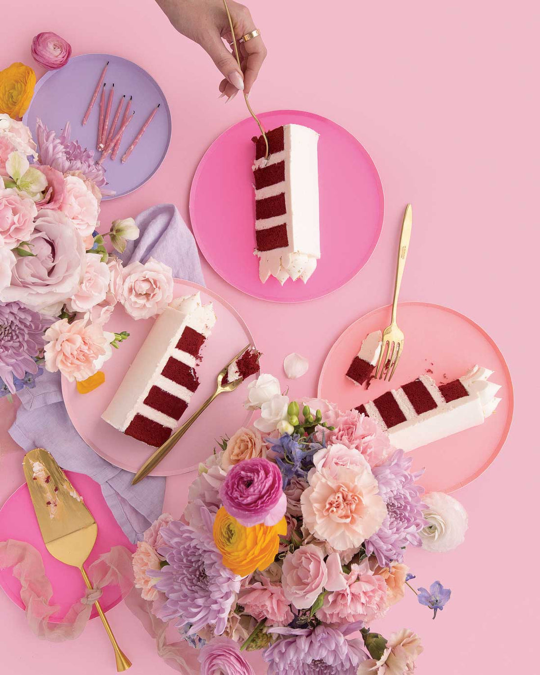 Three slices of Jenna Rae Cakes’ Red Velvet Cake with white frosting on pink plates, surrounded by pastel flowers and gold utensils, with a hand holding a fork above one slice, all set against a pink background.