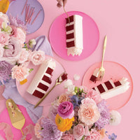 Three slices of Jenna Rae Cakes’ Red Velvet Cake with white frosting on pink plates, surrounded by pastel flowers and gold utensils, with a hand holding a fork above one slice, all set against a pink background.