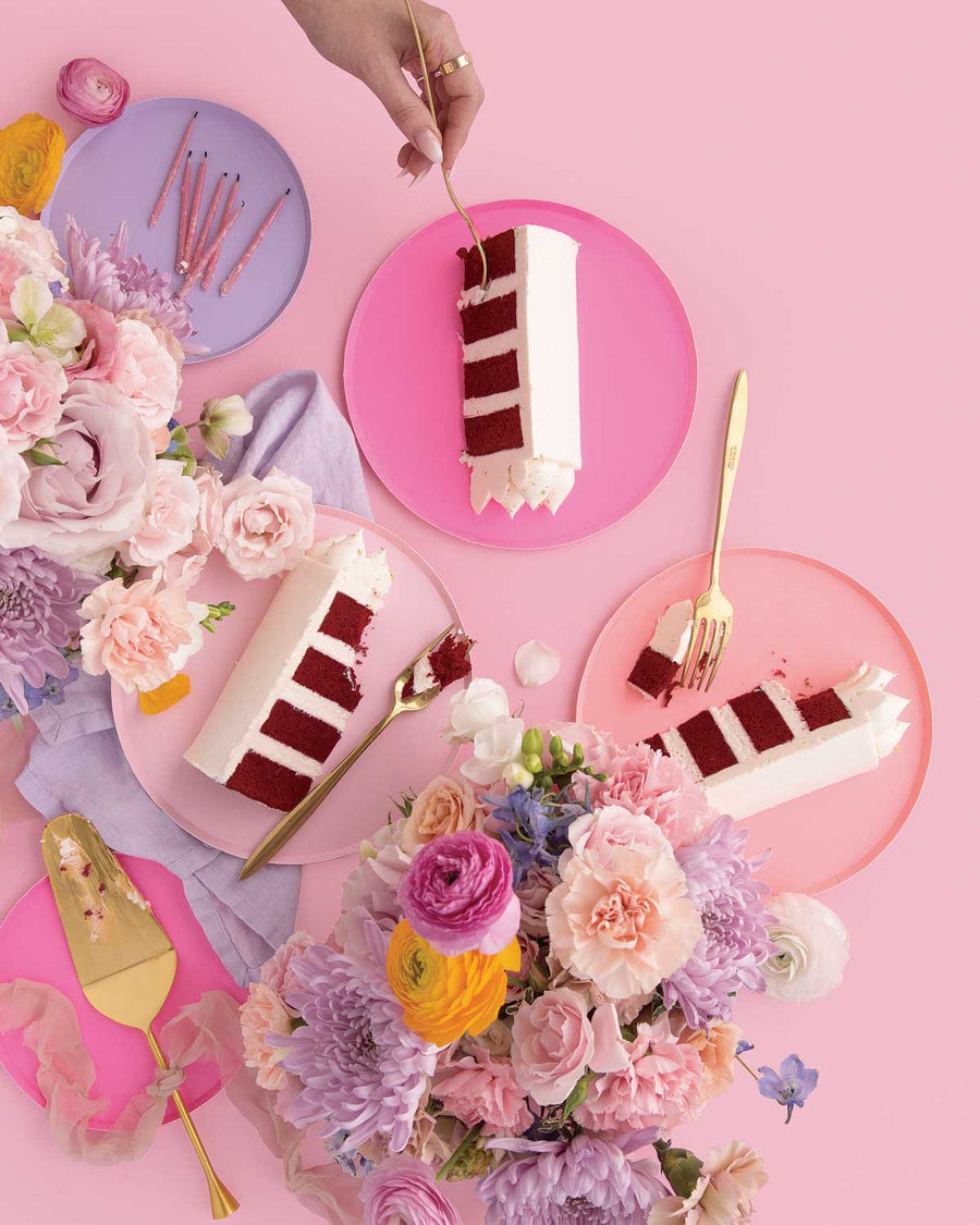 Three slices of Jenna Rae Cakes’ Red Velvet Cake with white frosting on pink plates, surrounded by pastel flowers and gold utensils, with a hand holding a fork above one slice, all set against a pink background.
