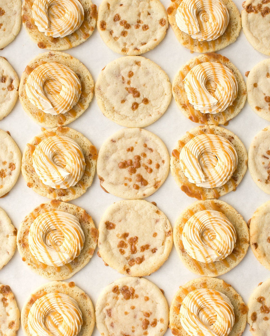 Jenna Rae Cakes Regular Cookie Sandwiches are displayed on parchment paper, featuring plain cookies, some with caramel bits, and others topped with swirled white and caramel-colored frosting.
