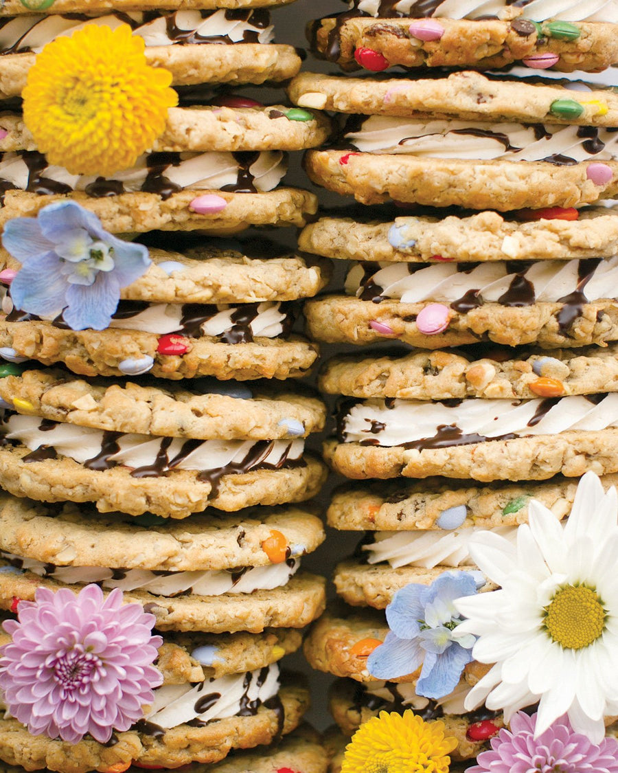 Regular Cookie Sandwiches by Jenna Rae Cakes are oatmeal cookies with colorful candies, white cream filling, and a chocolate drizzle, finished with fresh flowers like daisies and pink or blue blossoms.