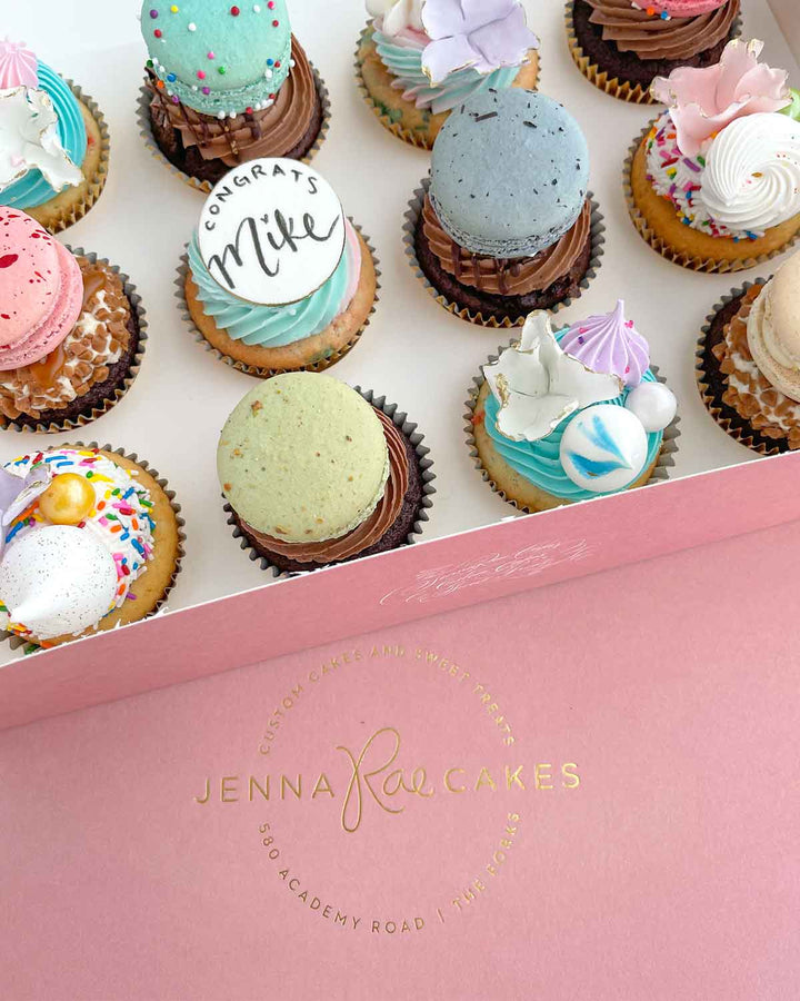 An open pink Jenna Rae Cakes box displays The Works Cupcakes, each decorated with colorful macarons, meringues, sprinkles, and a cookie that reads “Congrats Mike.”.