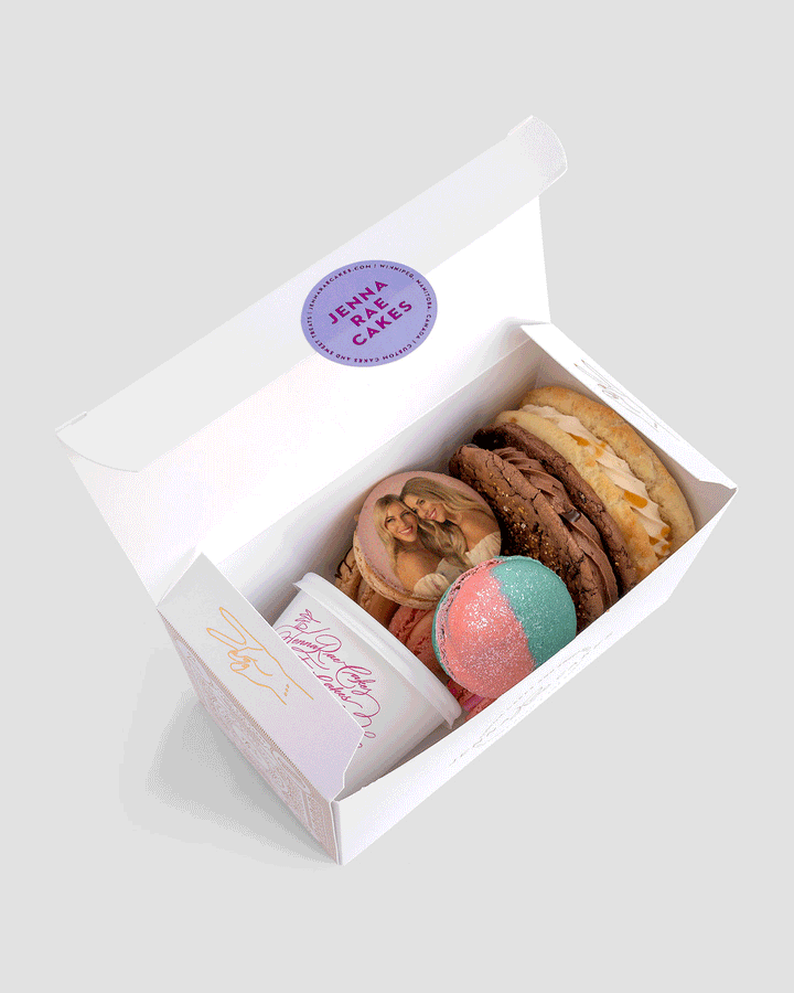 The Jenna Rae Cakes Sweet Treat Gift Box includes two large cookies, a pink and blue round treat, a small tub of frosting, and a macaron printed with two women smiling.