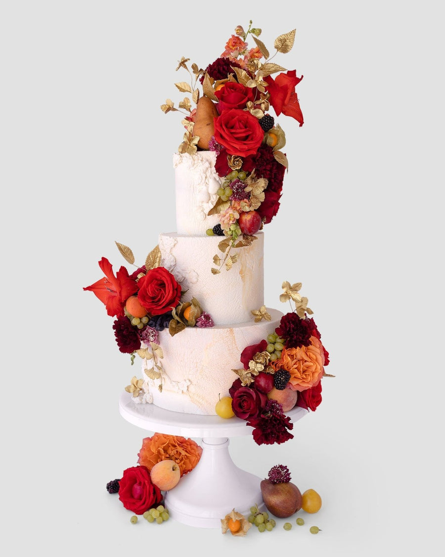 A two-tiered white cake on a stand, decorated with vibrant red and orange flowers, gold leaves, and assorted fresh fruits like figs, grapes, and berries, against a plain light gray background.