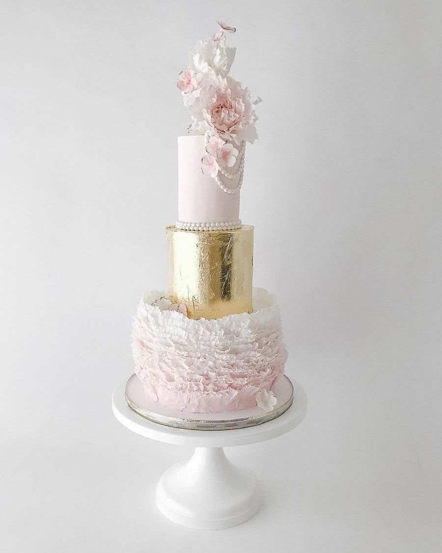 A three-tiered cake with pink, gold, and white layers; decorated with ruffled icing, gold leaf, pearls, and delicate pink and white sugar flowers, displayed on a white cake stand against a plain background.